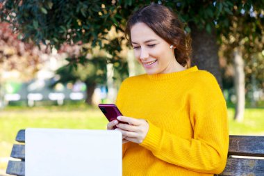 Dizüstü bilgisayar kullanan ve elinde akıllı telefon tutan genç bir kadın. Mutlu kadın parkta bir video konferans görüşmesi yapıyor. Bankta oturuyor.