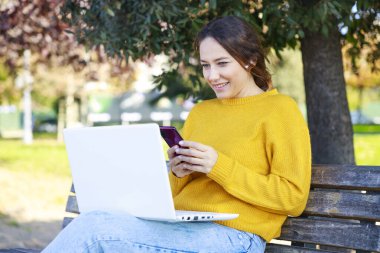 Dizüstü bilgisayar kullanan ve elinde akıllı telefon tutan genç bir kadın. Mutlu kadın parkta bir video konferans görüşmesi yapıyor. Bankta oturuyor.