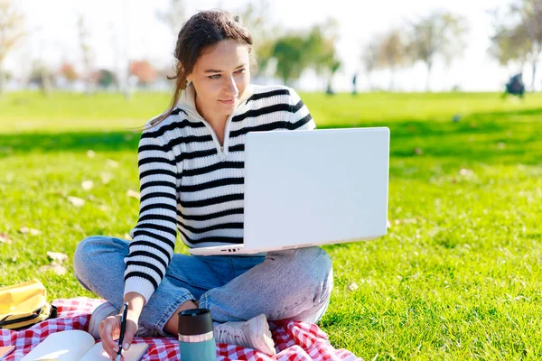 stock image Young woman sitting on grass at park with laptop and making notes in her diary. Freelancer female working while sitting at park.