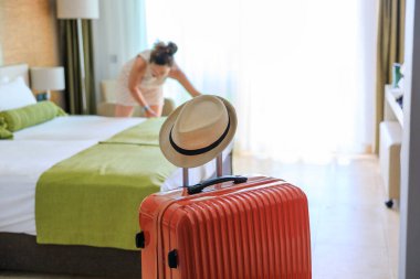 Otel odasında turuncu renkli bavul teslim edildi. Genç bir kadın yatak çarşaflarını değiştiriyor.