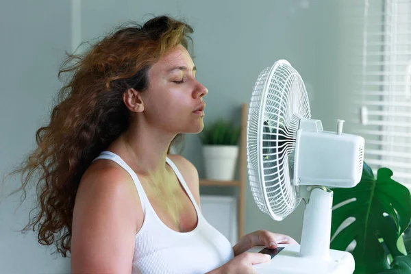 Young woman indoors, trying to cool off in front of an electric fan. She is standing by a window with her face close to the electric fan