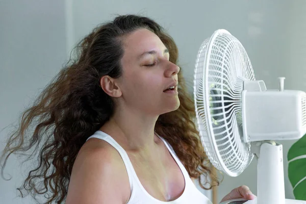 Young woman indoors, trying to cool off in front of an electric fan. She is standing by a window with her face close to the electric fan