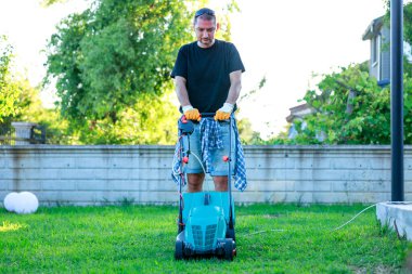 Güneşli bir günde arka bahçesinde çim biçme makinesiyle çim biçen genç adam.