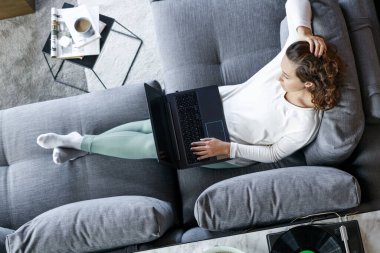 Evdeki kanepede dizüstü bilgisayarla oturan genç bir kadının en iyi görüntüsü.