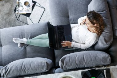 Evdeki kanepede dizüstü bilgisayarıyla oturan ve telefonla konuşan genç bir kadının en iyi görüntüsü.
