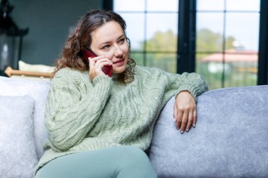 Evde telefonla konuşan mutlu genç bir kadın, elinde akıllı bir telefonla gülümseyen bir kadın, oturma odasında arama yapıyor.