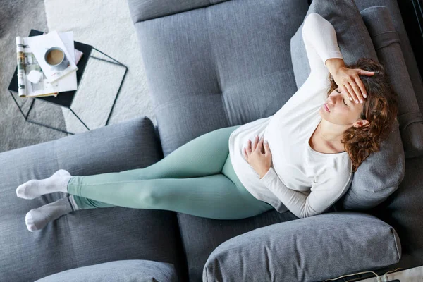 自宅でソファーで胃の痛みや頭痛を経験している若い女性の高角度ショット — ストック写真
