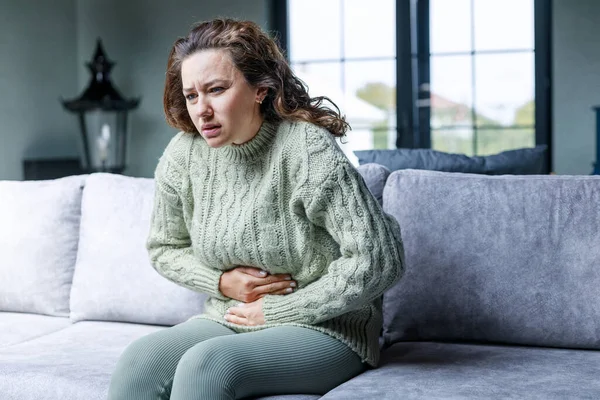 Sad young woman sitting on the couch and feeling spasm and symptoms of pms
