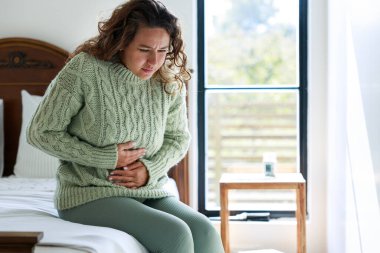 Sad young woman sitting on the bed and feeling spasm and symptoms of pms clipart