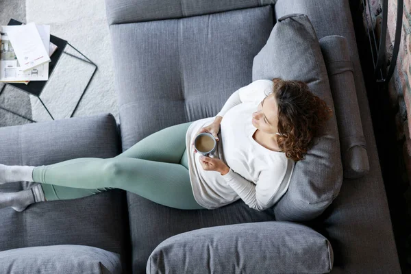 リビングルームのソファーに横たわる若い女性がコーヒーを汚し 耳のヘッドフォンから音楽を聴く — ストック写真