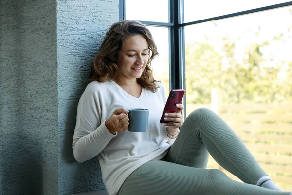 Młoda Kobieta Trzyma Filiżankę Kawy Używa Telefonu Siedząc Parapecie Domu — Zdjęcie stockowe