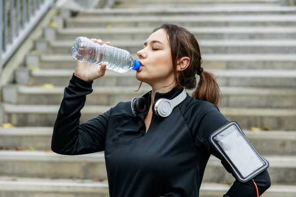 Young sportswoman drinking water from a bottle after workout on city background