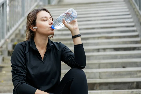 Young sportswoman drinking water from a bottle after workout on city background