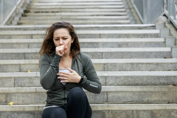 Young sporty woman coughing while exercising and sitting on staircase at street. Athlete woman affected by air pollution during running training