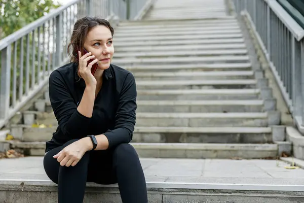 年轻的女人坐在楼梯上 在电话里说话 微笑的女运动员在城市里锻炼时休息一下 健身和健康生活方式概念 图库照片