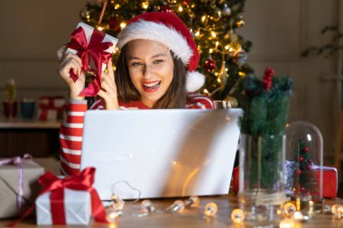 Neşeli kadın elinde bir hediye kutusu tutarken dizüstü bilgisayar kullanıyor ve Noel akşamında video görüşmesi yapıyor.