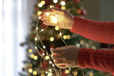 Bir kadının elleri parıldayan Noel ağacının önünde Noel ışıklarını tutuyor.