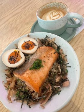 Üzerine yosun, bonito taneleri, kaynamış yumurta ve yeşil soğan seramik kupada latte sanatıyla birlikte ızgara somon balığı kasesi. Sıcak kafe atmosferli ev yapımı öğle yemeği.