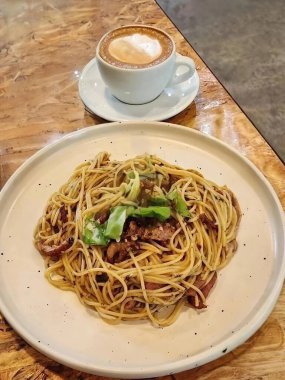 Taze yeşillik ve kızarmış pastırmayla süslenmiş spagetti, yanında sıcak kapuçino ile servis ediliyor. Kafeterya sahneleri ya da yemek görselleri için mükemmel, rahat, tatmin edici bir yemek..