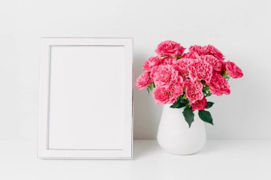 Beyaz çerçeveli maket ve pembe gül buketi beyaz bir vazoda, duvardaki beyaz bir masada. Uzayı metin için kopyala. Tatil kavramı