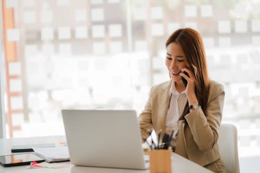 Genç Asyalı iş kadını müşterilerle konuşmak için akıllı telefon kullanıyor, hesaplamak için laptop ve finansal grafiklerle çalışıyor, mali muhasebe, çevrimiçi iş konsepti.