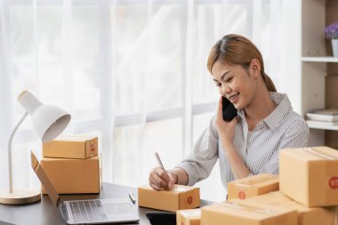Gülümseyen Asyalı kadın kameraya bakıyor ve müşteriye göndermeden önce paket teslimat bilgilerini kontrol etmek için dizüstü bilgisayar kullanıyor. Küçük çaplı bir girişimci. Çevrimiçi satış kavramı.
