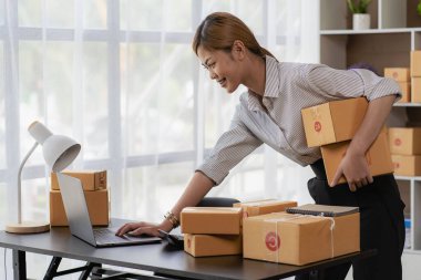 Gülümseyen Asyalı kadın kameraya bakıyor ve müşteriye göndermeden önce paket teslimat bilgilerini kontrol etmek için dizüstü bilgisayar kullanıyor. Küçük çaplı bir girişimci. Çevrimiçi satış kavramı.