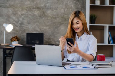 Akıllı telefonu elinde tutan Asyalı kadın muhasebeci masadaki grafik dokümanlarıyla finansal hesap verilerini analiz ediyor..