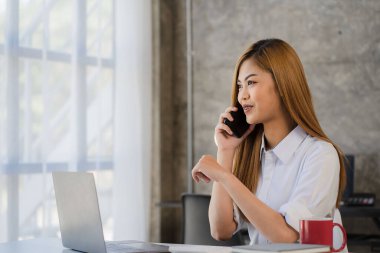 Akıllı telefonu elinde tutan Asyalı kadın muhasebeci masadaki grafik dokümanlarıyla finansal hesap verilerini analiz ediyor..