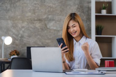 Akıllı telefonu elinde tutan Asyalı kadın muhasebeci masadaki grafik dokümanlarıyla finansal hesap verilerini analiz ediyor..