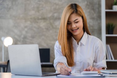 Finansal muhasebede çalışan Asyalı bir kadın hesap makinesi kullanarak çizelgeleri analiz ediyor, belgeli dizüstü bilgisayarı kullanıyor ve elinde kalemlerle finans bürosundaki bir masada oturuyor..
