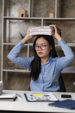 Asyalı kadın işçi gece geç saatlere kadar ofiste çalışıyor kız çok fazla çalışıyor ve masa başında kağıt yığınlarıyla çalışırken stres ve baş ağrısı sorunu yaşıyor..