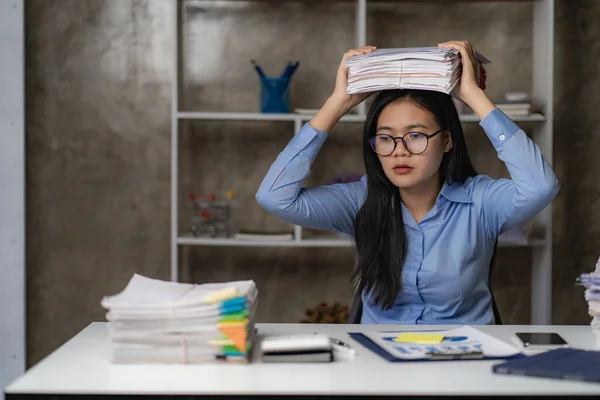 夜遅くにオフィスで残業しているアジアの女性労働者女の子は過労しており 机の上の書類の山で作業中にストレスや頭痛を感じるのに苦労しています — ストック写真