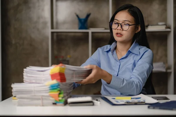 Asian female worker working overtime in the office late at night The girl is overworked and has trouble with feeling stress and headache while working with piles of papers on the desk.
