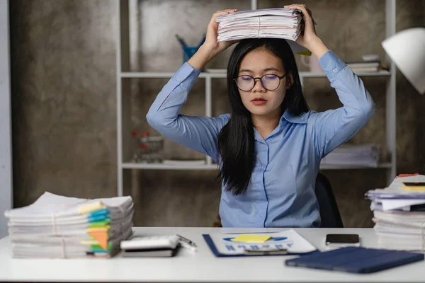 夜遅くにオフィスで残業しているアジアの女性労働者女の子は過労しており 机の上の書類の山で作業中にストレスや頭痛を感じるのに苦労しています — ストック写真