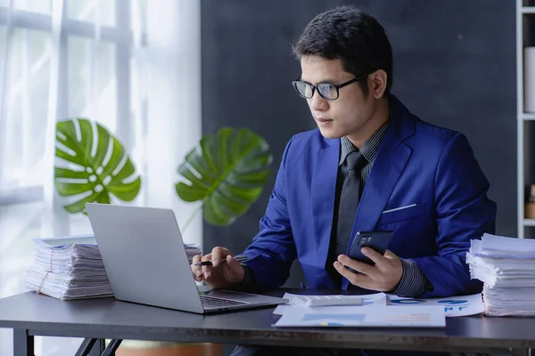 Asian business man wearing glasses in a blue suit working with piles of financial documents on the table, financial analysis concept.