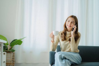 Akıllı telefon kullanan Asyalı kadın evde kanepede otururken mutlu bir şekilde gülümseyen sosyal medyayı kontrol ediyor.