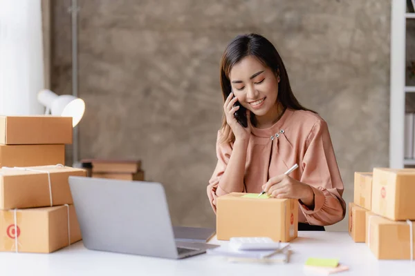 stock image An Asian woman starts a small independent business sitting with parcel boxes and laptops, three smartphones on a wooden table in her living room at home.