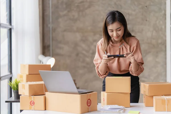 stock image An Asian woman starts a small independent business sitting with parcel boxes and laptops, three smartphones on a wooden table in her living room at home.