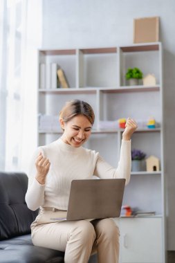 Kanepede oturan gülümseyen Asyalı kadın evdeki oturma odasında ev bilgisayarı kullanıyor. Mutlu kız öğrenciler rahatlamak, çalışmak ve çalışmak için bilgisayar kullanırlar..
