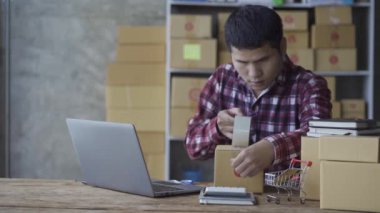 Akıllı telefondan konuşan ve evde online ürün satmak için dizüstü bilgisayar kullanan genç adam. Satış elemanı, mağazadan sipariş veren müşterilere teslim etmek için ürünleri kutulara koyar. Satıcı, alışveriş sitesi, işyeri sahibi, küçük KOBİ iş fikri.