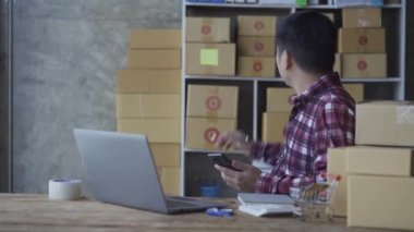 Akıllı telefondan konuşan ve evde online ürün satmak için dizüstü bilgisayar kullanan genç adam. Satış elemanı, mağazadan sipariş veren müşterilere teslim etmek için ürünleri kutulara koyar. Satıcı, alışveriş sitesi, işyeri sahibi, küçük KOBİ iş fikri.