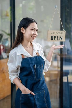 Asyalı bir KOBİ sahibi önlük takıyor ve açık tabelası olan bir kahve dükkanının kapısında duruyor. Asyalı iş kadını Kafe sahibi, barista, KME girişimcisi, satıcı konsepti
