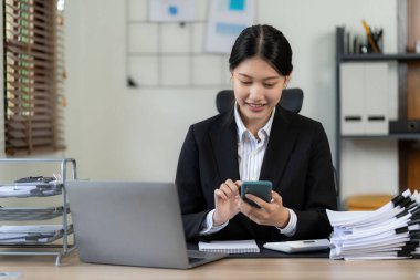 Telefonunu, bilgisayarını ve finansal belgelerini masasında kullanırken gülümseyen bir yüzü olan genç Asyalı kadın muhasebeci. Konseptleri planlıyor, pazar verilerini analiz ediyor..