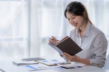 Masandaki dizüstü bilgisayarla pazarlama verilerini planlayan ve analiz eden bir yığın finansal grafik çizelgesi ile çalışan genç Asyalı kadın. Şirket bilgi bürosu