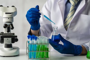 Laboratuvar önlüklü bir bilim adamı ya da doktor, biyokimyasal laboratuvar araştırması yapan cam bir aparatta reaktif mikroskop ve renkli bir sıvı kullanarak test tüpü tutuyor..