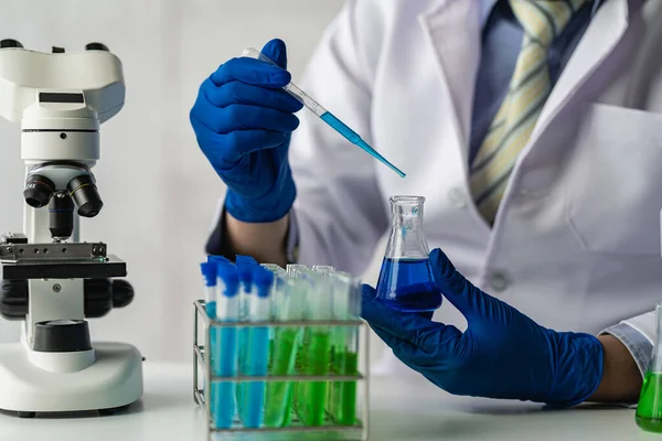 stock image A scientist or a doctor in a lab coat holds a test tube using a reagent microscope and a drop of colored liquid on a glass apparatus doing biochemical laboratory research.