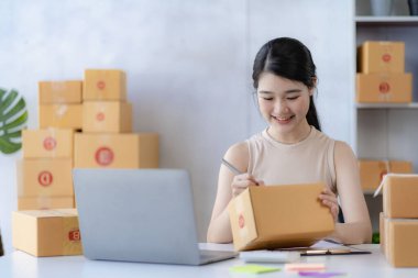 Genç Asyalı kadın internette KOBİ işi için bir kutu ve dizüstü bilgisayar satarak çalışıyor. Paket teslim kavramı.