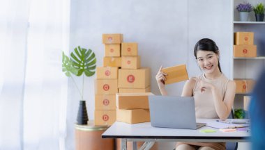 Genç Asyalı kadın internette KOBİ işi için bir kutu ve dizüstü bilgisayar satarak çalışıyor. Paket teslim kavramı.