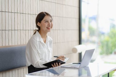 Çekici Asyalı kadın kafede bir masada tek başına oturuyor iş için dizüstü bilgisayarına bakıyor gibi görünüyor ve gülümsüyor, internette çalışıyor gibi..